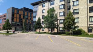 The image shows the exterior two large square modern buildings each about 5 stories high. The street in front of the buildings, including angled parking spots, is prominent in the foreground of the image. 