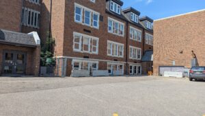 The image shows an accessible parking spot, which is located among other parking stalls, at the exterior of a large brick building with many windows. 