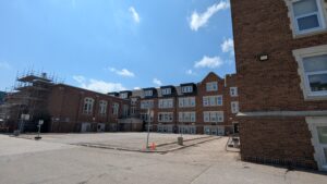 The image shows a small square parking lot that is located at the exterior of a large older brick building with many windows.