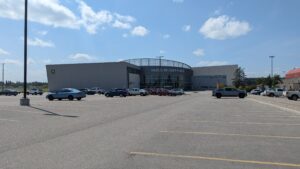 This image is dominated by a large paved parking lot in the foreground. There is a large modern building with a partial circular exterior in the background, and blue skies with clouds above. 