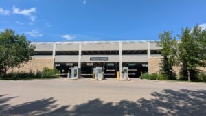 The image shows a blocky, concrete 3-level parkade, with a shadowed street in front, and blue sky overhead.