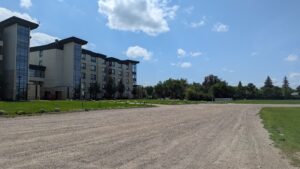 The image predominantly shows a gravel road and parking lot, with a large, modern square buidling, trees, and a big blue sky with clouds in the background. 